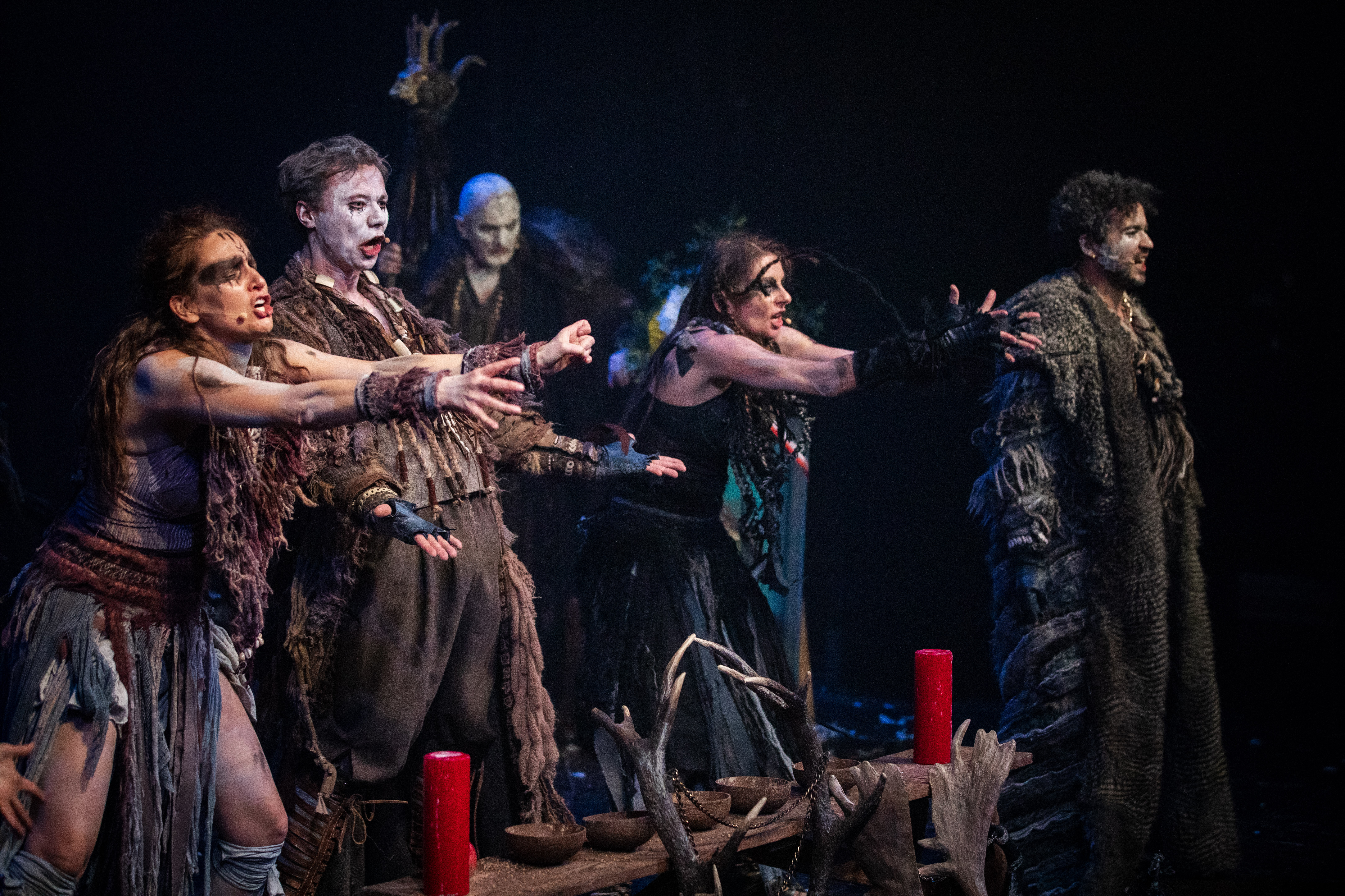 5 aktorów w kostiumach ustawionych w rzędzie na scenie, śpiewają i wyciągają ramiona przed sobą